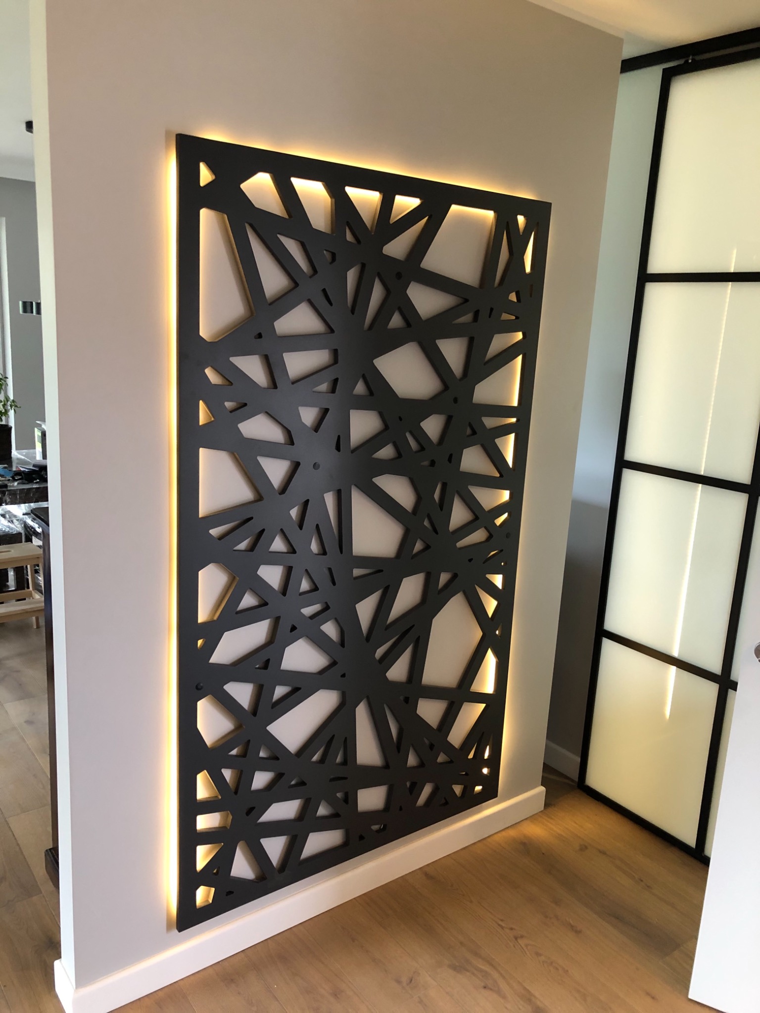 Dekoracyjny panel ścienny z podświetleniem LED w nowoczesnym wnętrzu, abstrakcyjny wzór geometryczny, ciemny kolor panelu na jasnej ścianie.