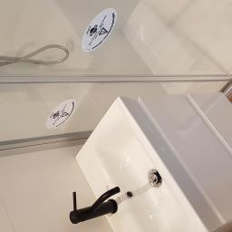 Szymon Zielinski - Biała umywalka z czarną baterią, kabina prysznicowa z naklejkami ostrzegawczymi, jasne płytki na ścianach i podłodze, imitacja drewna na podłodze w tle, widok z góry.