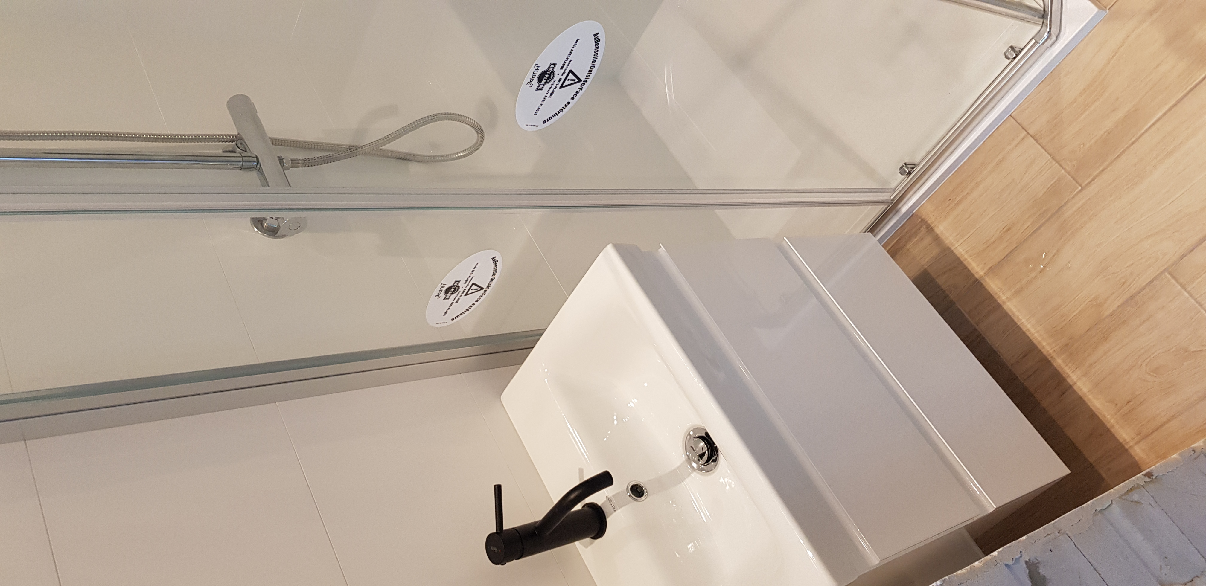 Biała umywalka z czarną baterią, kabina prysznicowa z naklejkami ostrzegawczymi, jasne płytki na ścianach i podłodze, imitacja drewna na podłodze w tle, widok z góry.