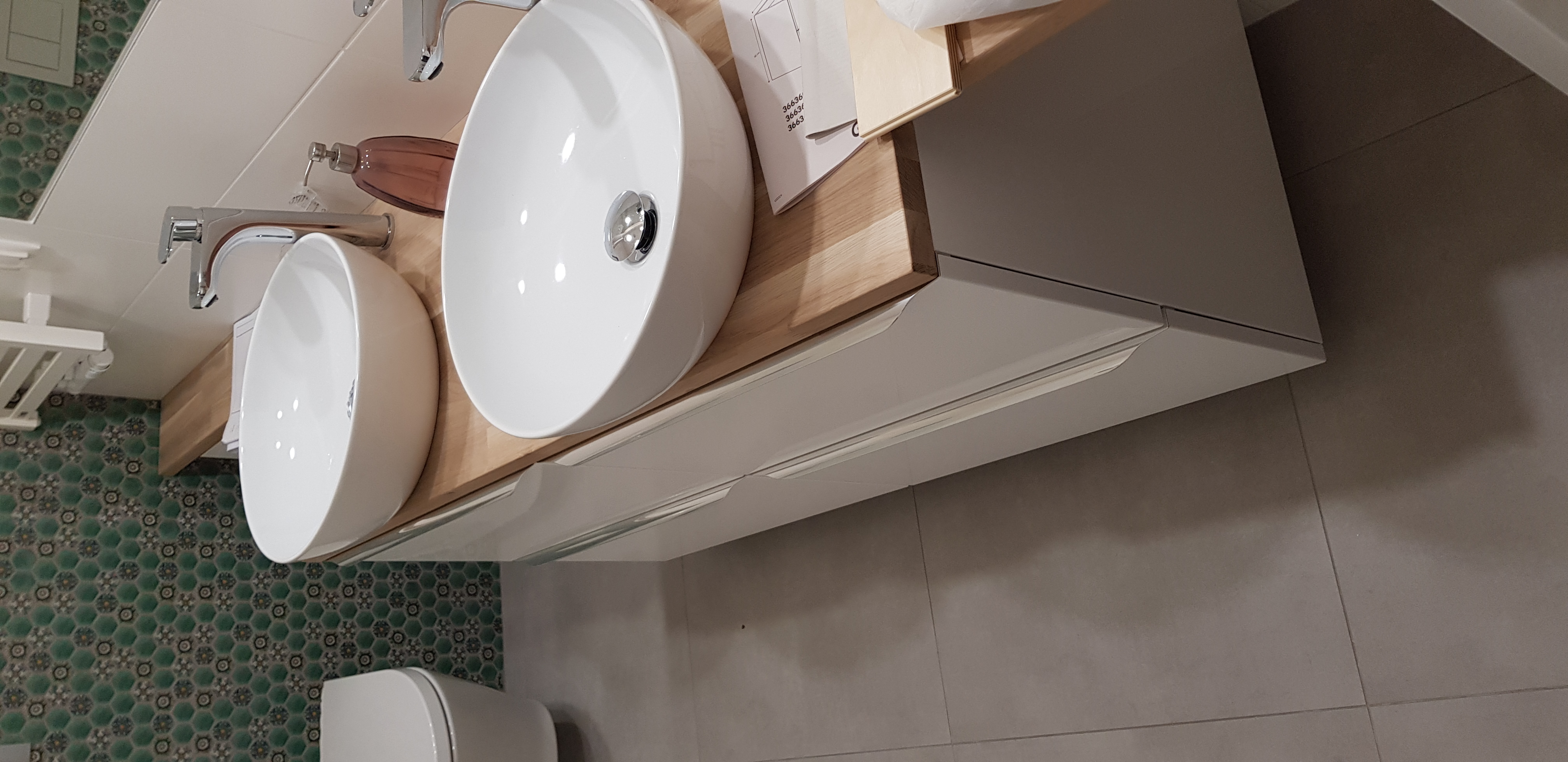 Dwie białe umywalki nablatowe o zaokrąglonym kształcie z chromowanymi bateriami, zamontowane na drewnianym blacie z szufladami w łazience z zieloną mozaiką na ścianie.