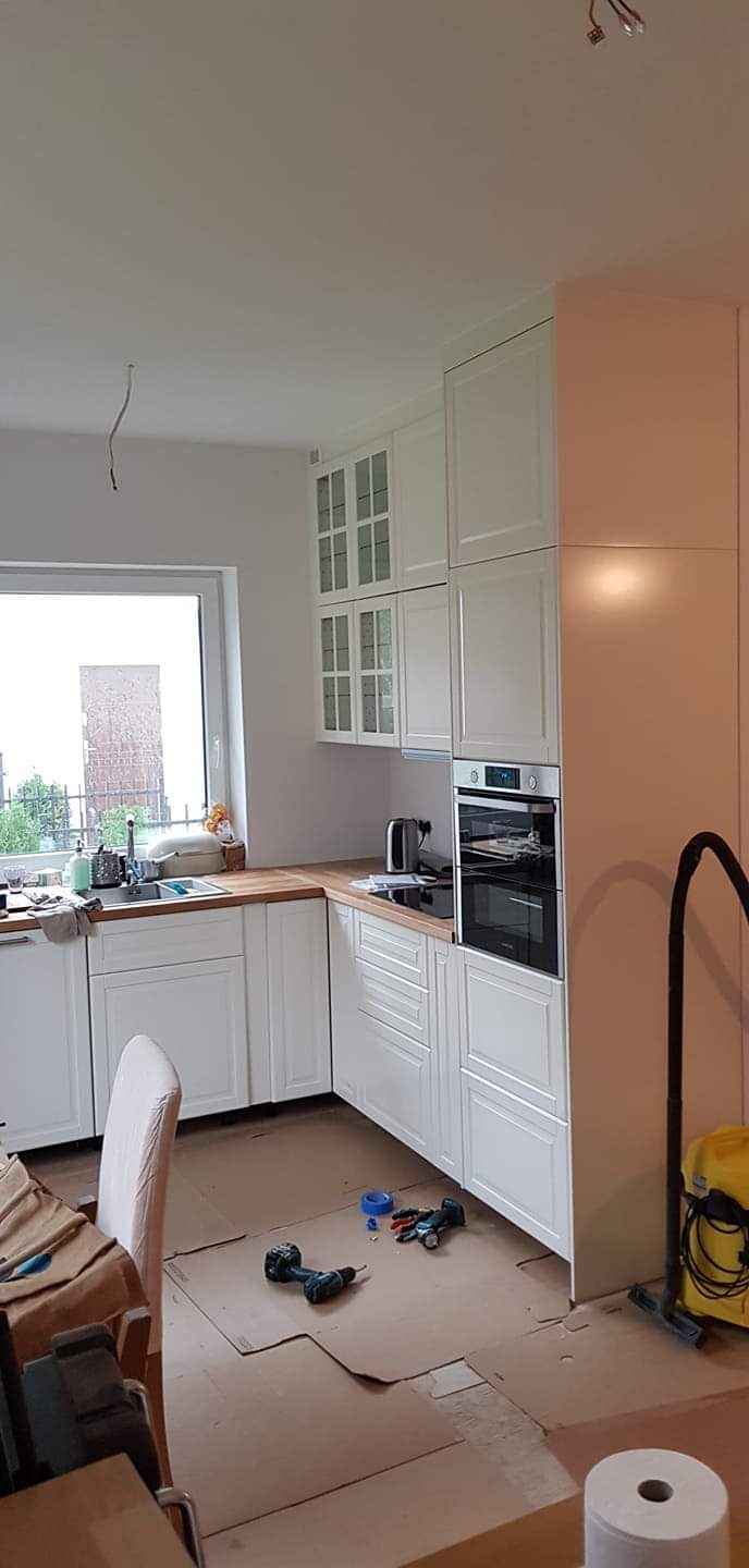 Wnętrze kuchni w trakcie montażu zabudowy meblowej: białe szafki z drewnianym blatem, widoczny piekarnik, okno z widokiem na zieleń, podłoga zabezpieczona kartonem, narzędzia i odkurzacz w tle.