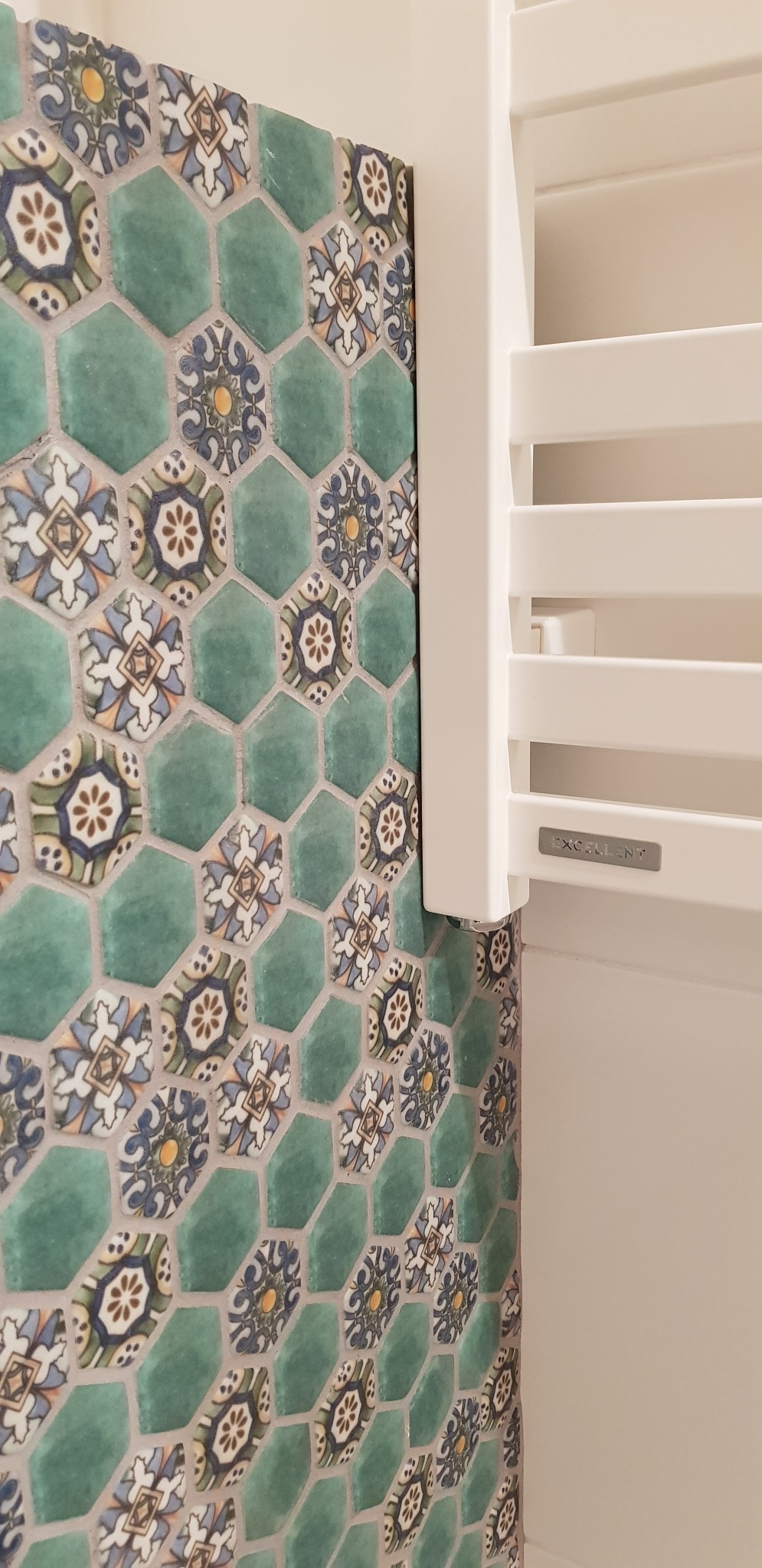 Fragment ściany wyłożonej heksagonalnymi, zielonymi płytkami z dekoracyjnym wzorem kwiatowym, sąsiadującej z białym, drabinkowym grzejnikiem łazienkowym marki Excellent.