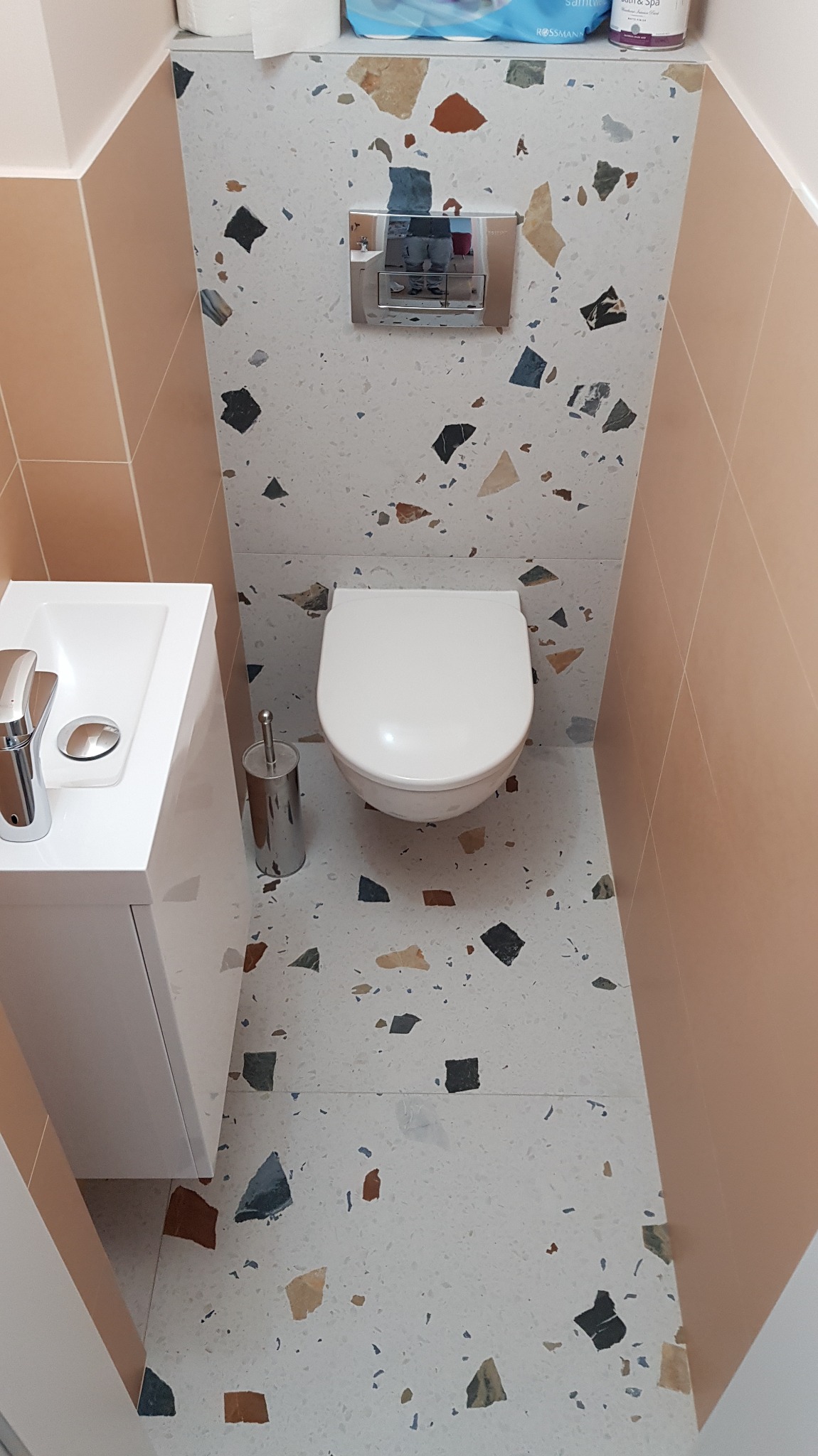 Wąska toaleta z umywalką, wykończona płytkami imitującymi lastryko na podłodze i ścianie za muszlą, kontrastująca z beżowymi płytkami na pozostałych ścianach.