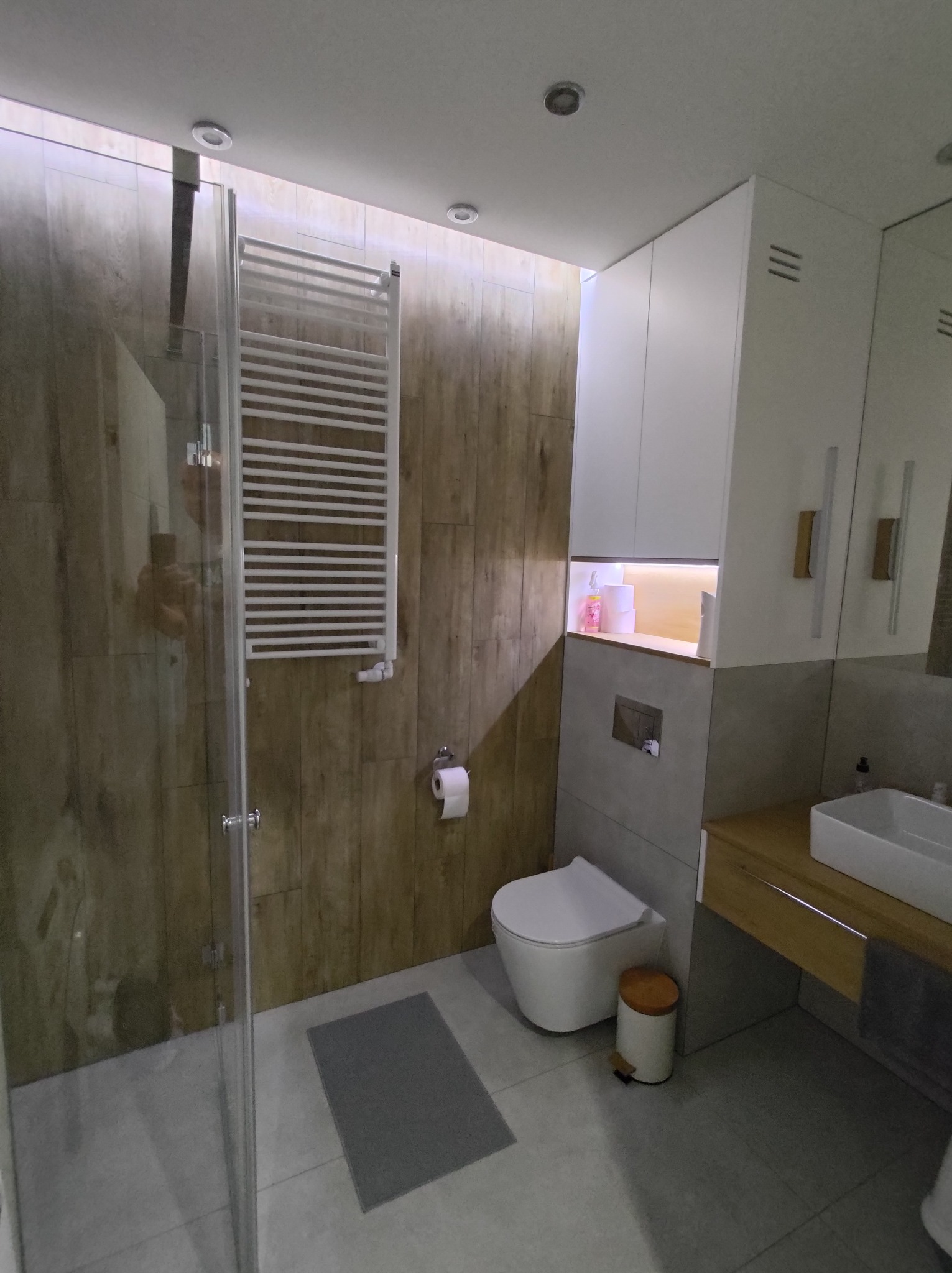 Nowoczesna łazienka z kabiną prysznicową ze szklanymi drzwiami, grzejnikiem drabinkowym na ścianie z imitacją drewna, wiszącą toaletą i umywalką nablatową na drewnianym blacie.