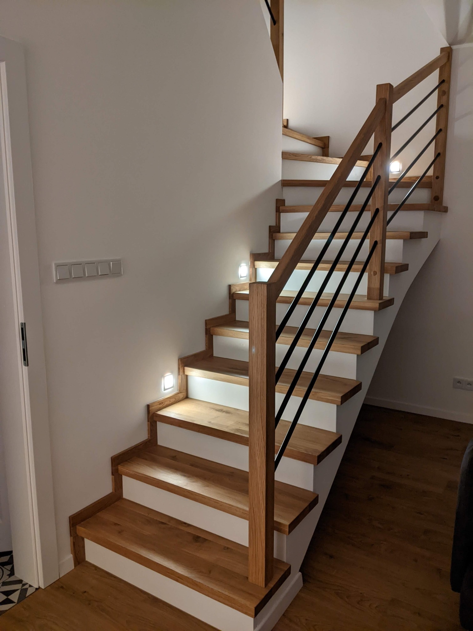 Drewniane schody z białymi podstopnicami i czarnymi metalowymi prętami balustrady, oświetlone punktowymi lampkami LED w ścianie.
