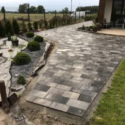 Świeżo ułożony, szary chodnik z kostki brukowej prowadzący do domu, z widocznymi elementami ogrodu: krzewami, drzewkami i białym żwirem. Na pierwszym planie wbita łopata, a w tle zaparkowany bus.