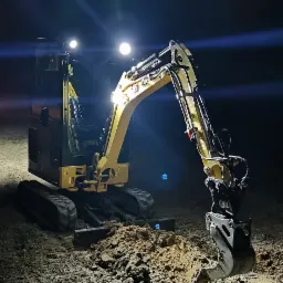 Żółta mini koparka oświetlona reflektorami podczas nocnych prac ziemnych, widoczne świeżo wykopane rowy.