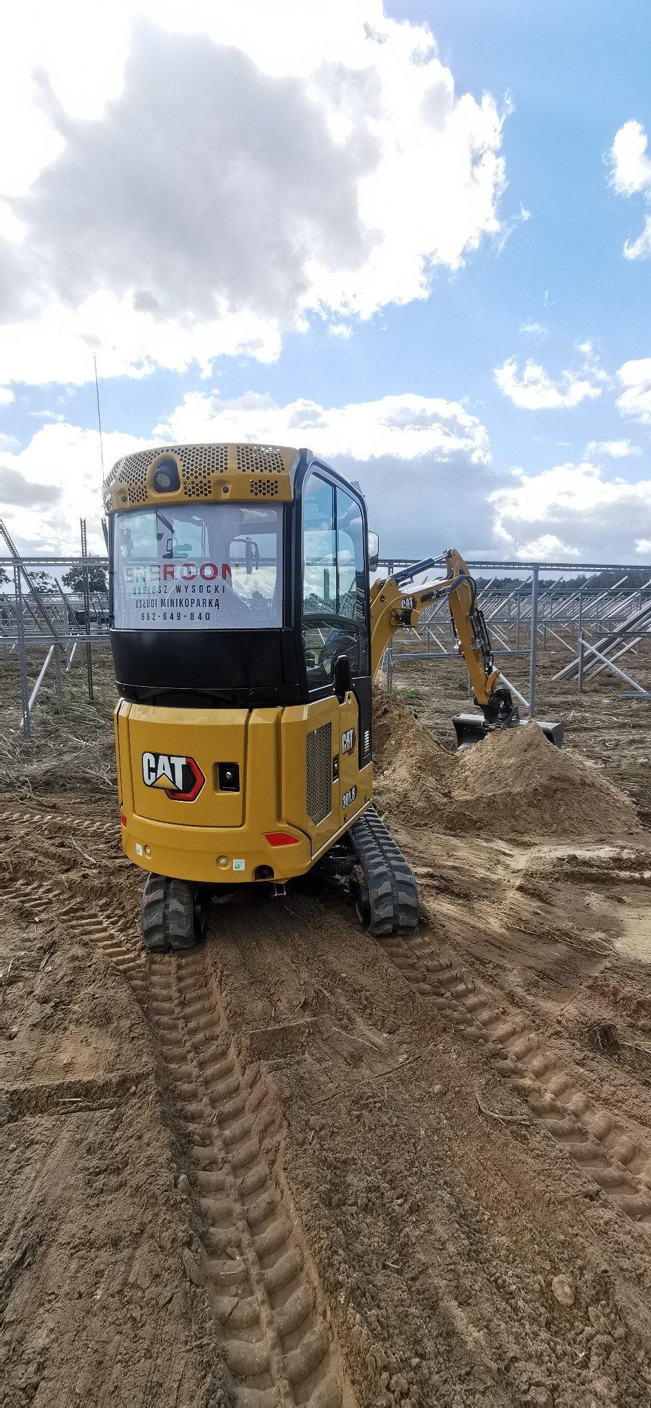 Żółta mini koparka CAT 301.8 z kabiną i naklejką firmy ENERCON na szybie, pracująca na placu budowy z elementami konstrukcji w tle, widoczne ślady gąsienic na ziemi.