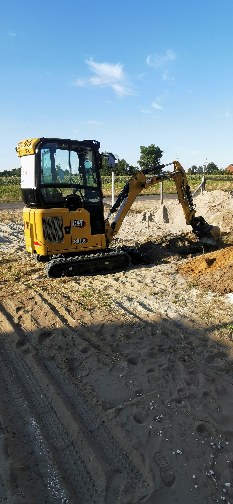 Żółta minikoparka CAT 301.8 w trakcie prac ziemnych, widoczne świeżo wykopane zagłębienie w piaszczystym podłożu, na tle błękitnego nieba i zieleni.