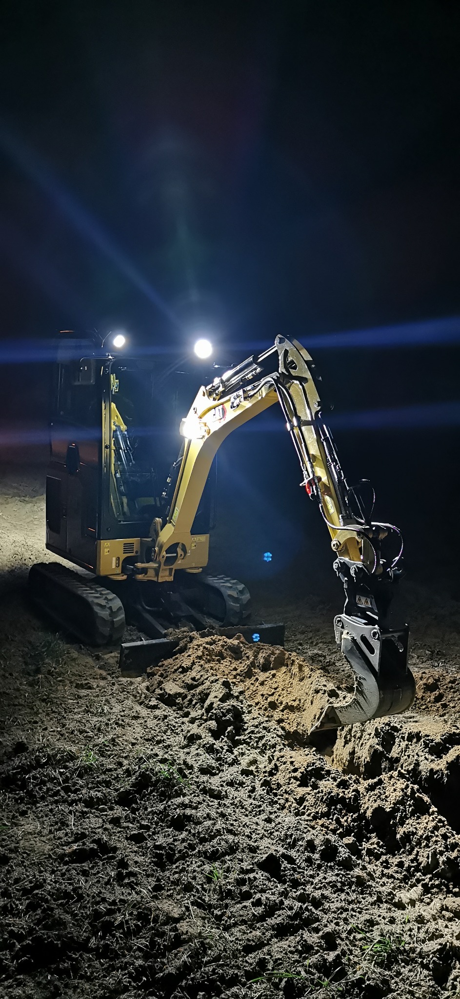 Żółta mini koparka oświetlona reflektorami podczas nocnych prac ziemnych, widoczne świeżo wykopane rowy.