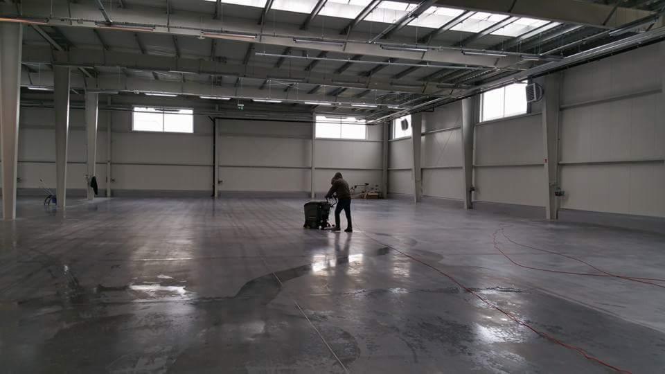 Pracownik w kombinezonie czyści przemysłową posadzkę w hali za pomocą szorowarki, widoczne ślady mokrej powierzchni i kable na podłodze.