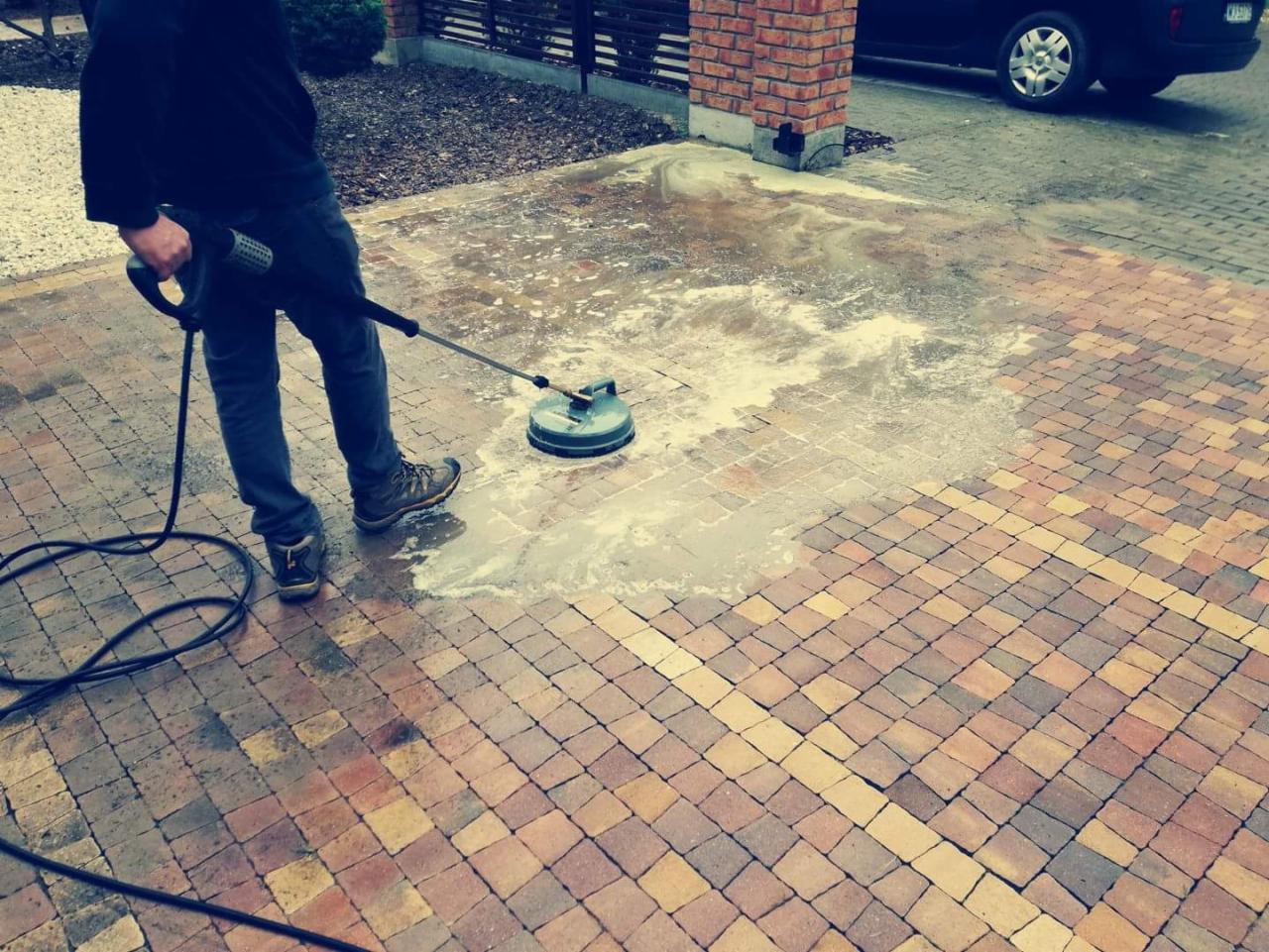 Czyszczenie kostki brukowej myjką ciśnieniową z rotacyjną dyszą, widoczna różnica między czystą a brudną powierzchnią, fragment muru z cegły klinkierowej w tle.
