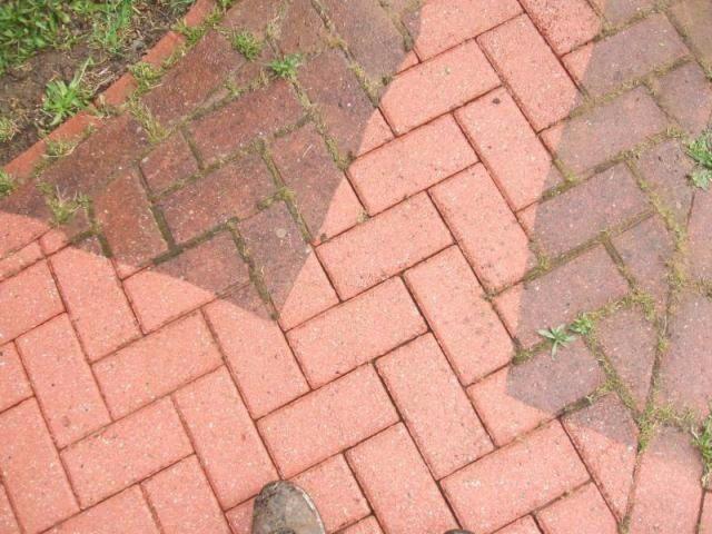 Częściowo wyczyszczona czerwona kostka brukowa ułożona w jodełkę, z widoczną różnicą między brudną a czystą powierzchnią oraz fragment buta w dolnym rogu.