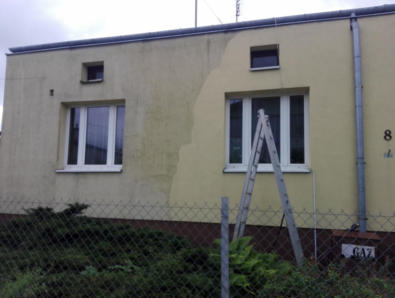 Elewacja budynku z widocznym efektem czyszczenia, część ściany umyta, a część brudna, drabina oparta o ścianę, krzewy i siatka w tle.