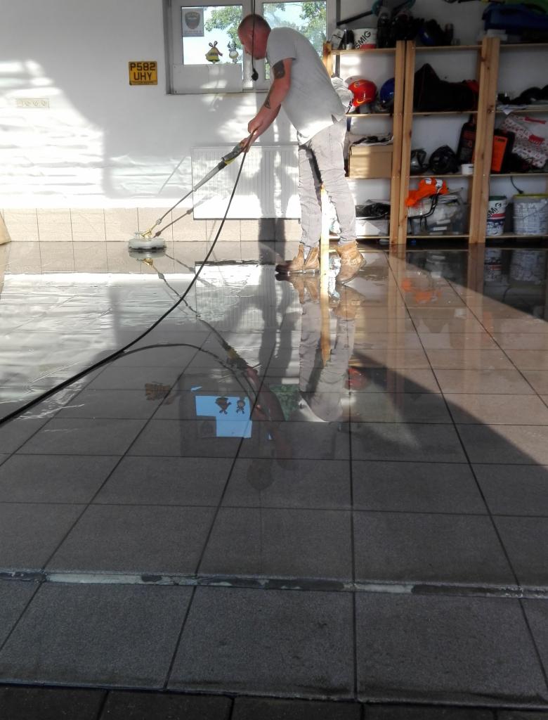 Mężczyzna w jasnych ubraniach czyści posadzkę z szarych płytek w garażu za pomocą elektrycznej szczotki, widoczne odbicie postaci w mokrej powierzchni, na ścianie żółta tablica rejestracyjna.