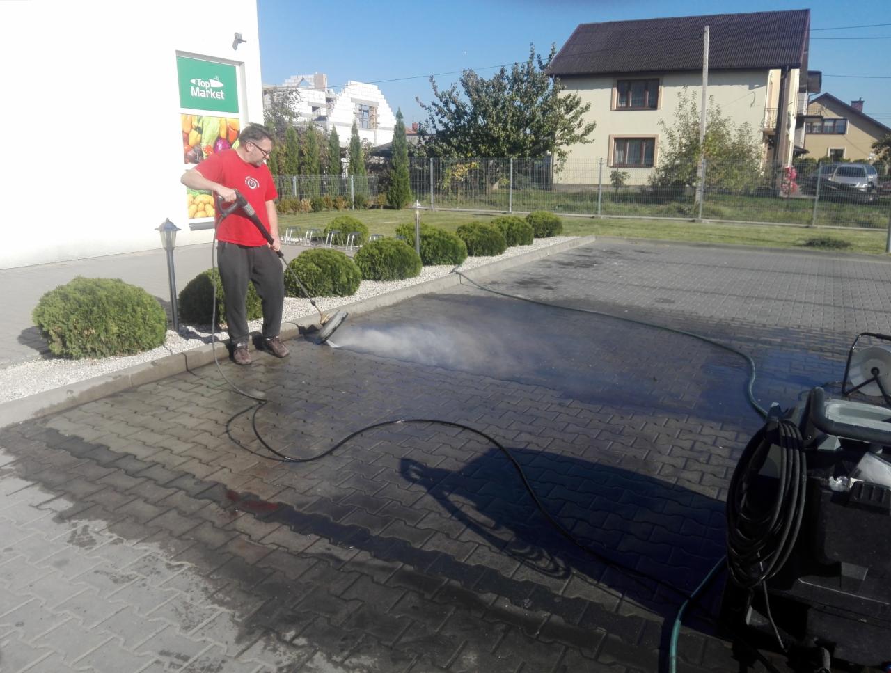 Pracownik w czerwonej koszulce czyści kostkę brukową przed sklepem Top Market za pomocą myjki ciśnieniowej, widoczne efekty czyszczenia.