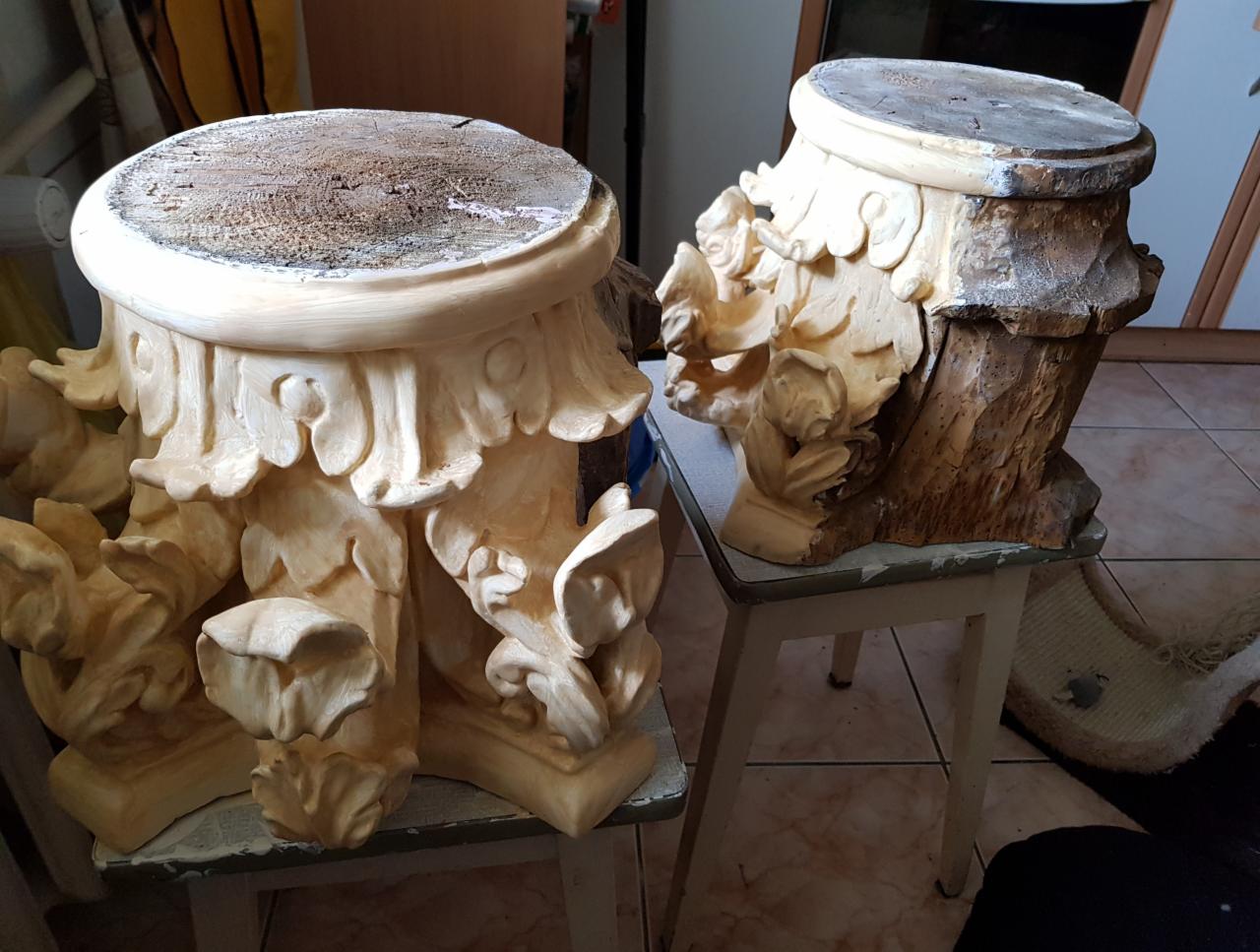 Dwa postumenty z drewna i gipsu, jeden z widoczną strukturą drewna, drugi z nałożoną warstwą gładzi i dekoracyjnymi elementami roślinnymi, oba stojące na starych krzesłach w warsztacie.