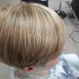 Krótka, prosta fryzura blond z widoczną strukturą włosów i delikatnym cieniowaniem, widok z góry, klientka w pelerynie ochronnej.