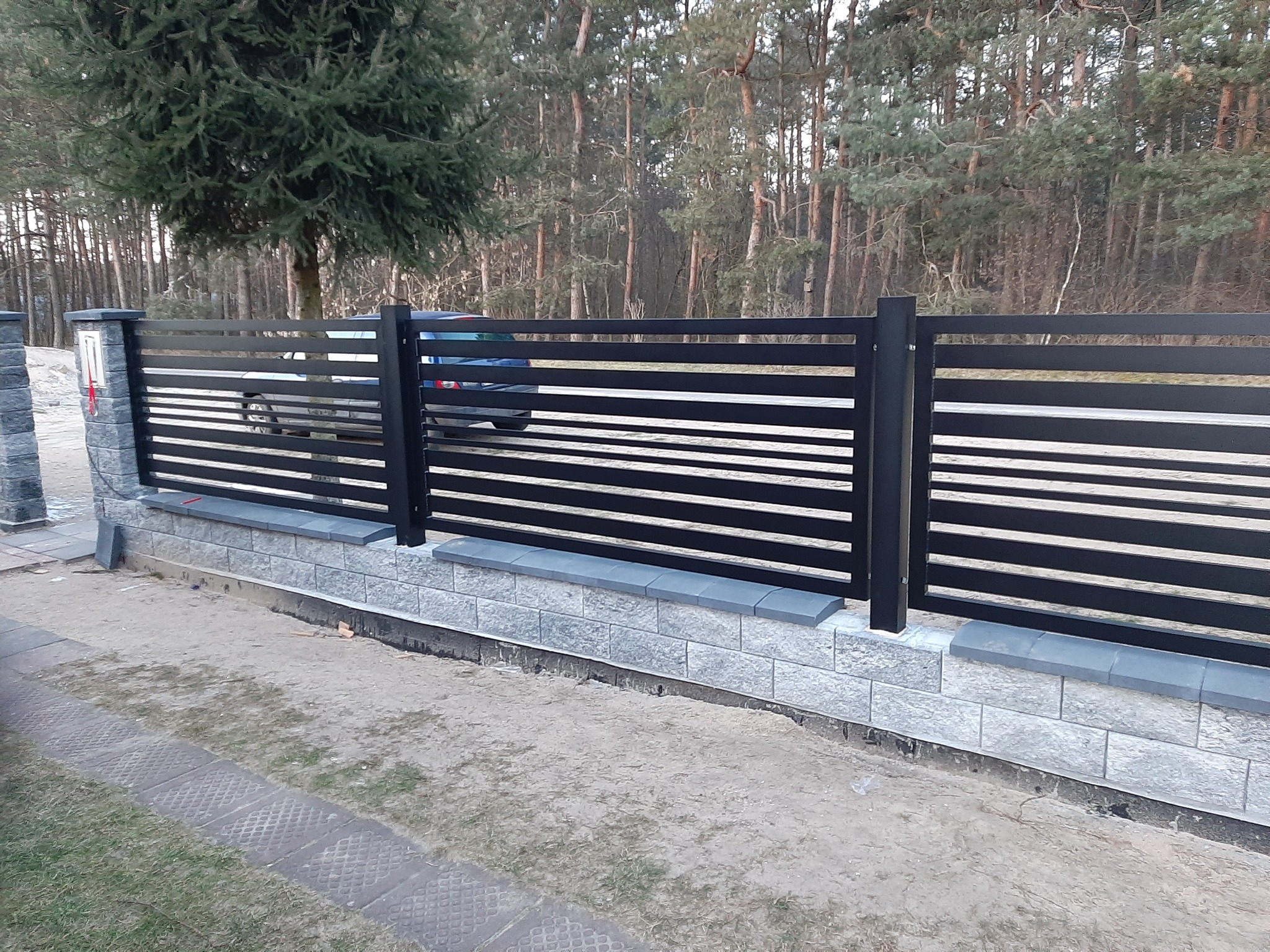 Nowoczesne ogrodzenie posesji z poziomymi, czarnymi panelami, osadzone na niskim murku z szarej kostki, w tle drzewa i fragment zaparkowanego samochodu.