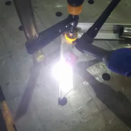 Zbliżenie na proces spawania TIG dw&oacute;ch metalowych element&oacute;w, z użyciem uchwytu spawalniczego z niebieską rękojeścią i zacisku śrubowego, generującego jasny łuk elektryczny na perforowanym stole...