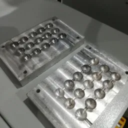 Dwie aluminiowe formy z wyżłobieniami w kształcie kulek połączonych cienkimi kanałami, leżące na szarym stole.