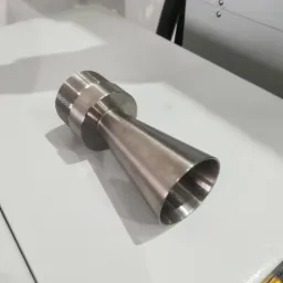 Metalowy element w kształcie stożka z gwintowanym zakończeniem, leżący na białym stole warsztatowym, widoczne ślady obróbki.