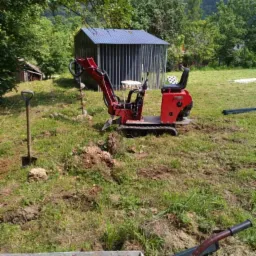 Czerwona mini wiertnica spalinowa wierci otwory pod słupki ogrodzeniowe na trawiastym terenie, widoczna łopata i taczka z betonem, w tle blaszany garaż z niebieskim dachem i zalesione wzgórze.