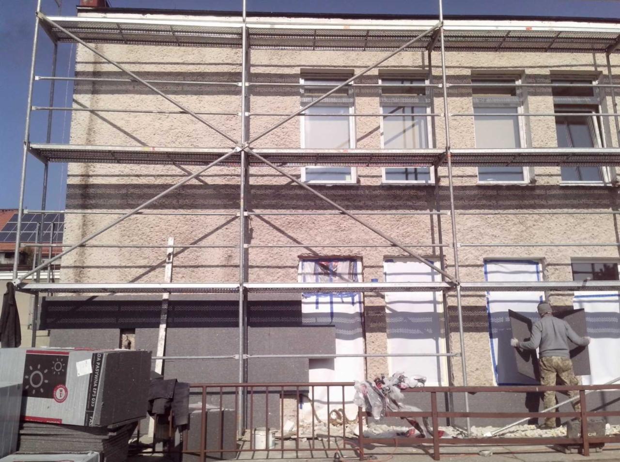 Pracownik na rusztowaniu przy ocieplaniu ściany budynku styropianem grafitowym, widoczne płyty styropianowe i materiały budowlane.