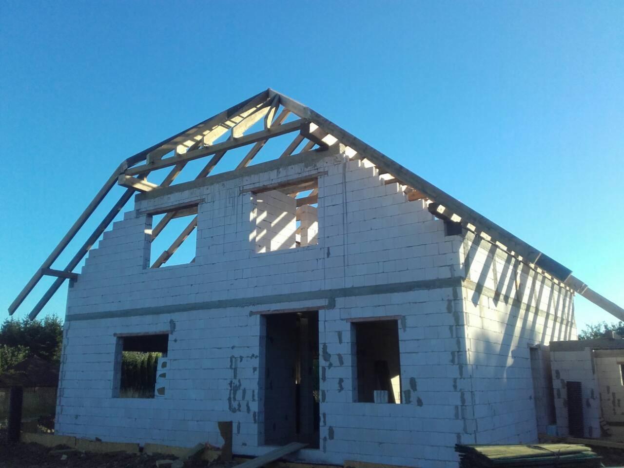 Wznoszenie ścian domu z bloczków betonowych, widoczne otwory okienne i drzwiowe, konstrukcja dachu w trakcie budowy, błękitne niebo w tle.