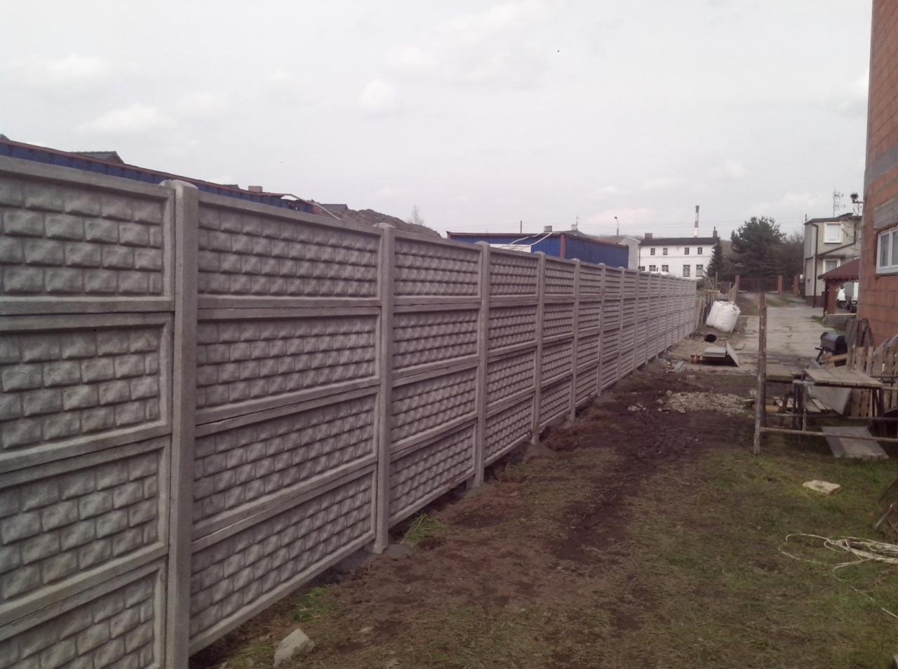 Długi, betonowy płot panelowy o wzorze cegieł oddziela posesję od drogi, widoczne budynki w tle, pochmurne niebo.