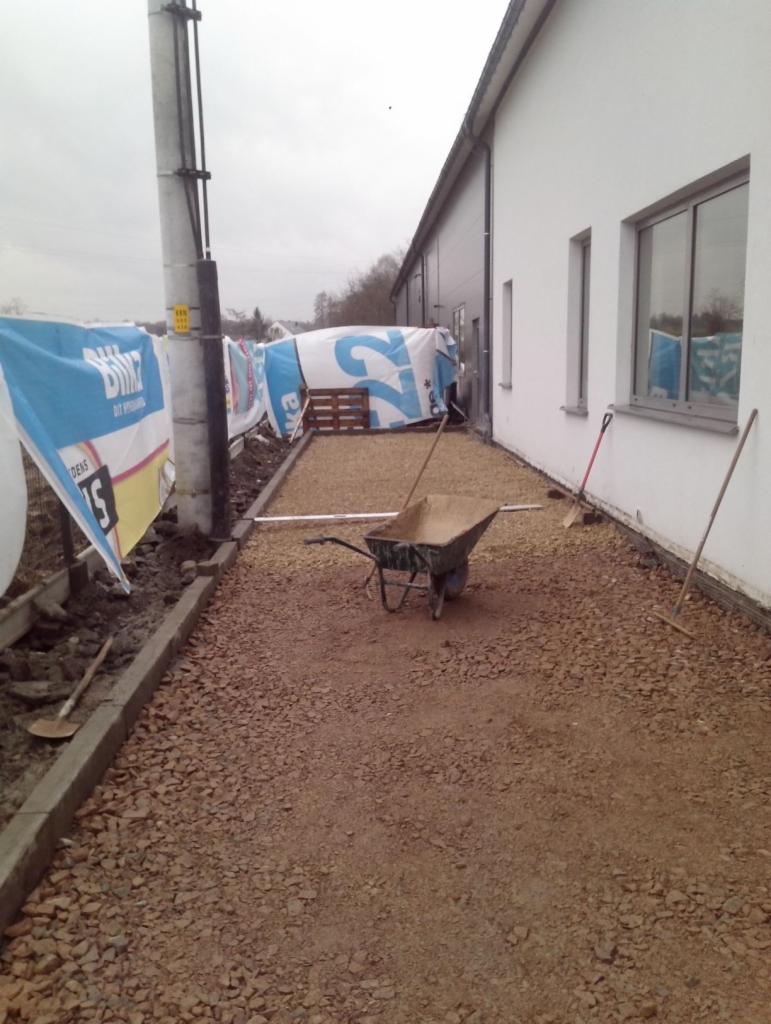 Przygotowanie podłoża z kruszywa pod ogrodzenie posesji, widoczna taczka, łopaty, krawężniki i materiały budowlane pod plandekami.