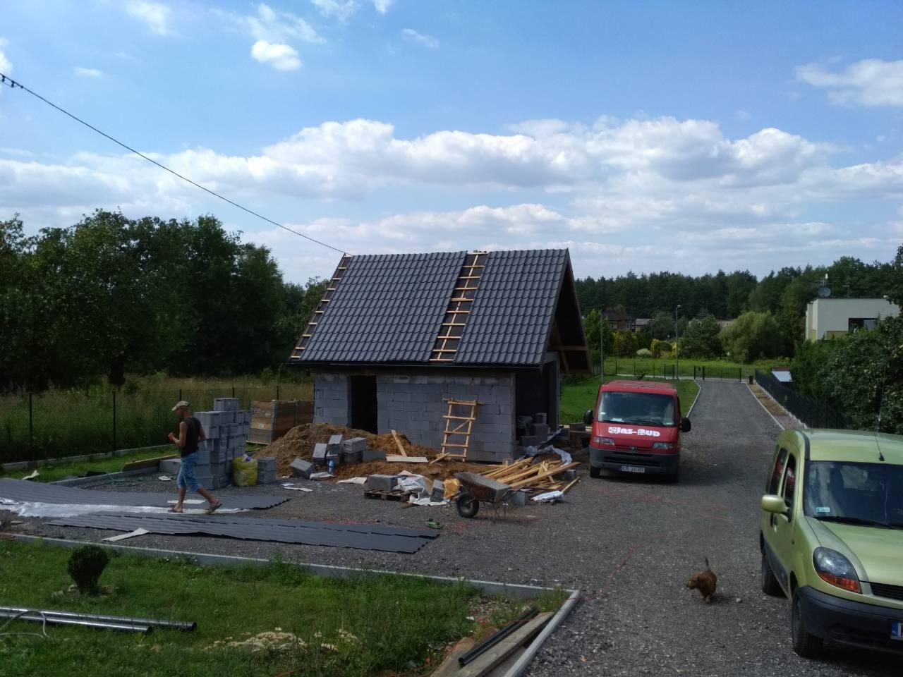 Budowa małego budynku gospodarczego z szarej cegły, widoczne materiały budowlane, drabina oparta o dach, pracownik w oddali, czerwony samochód dostawczy i zielony samochód osobowy na żwirowym...