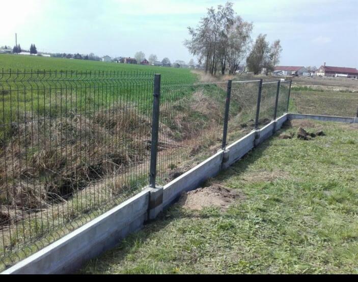 Panelowe ogrodzenie z podmurówką betonową na nierównym terenie, widok na pole i zabudowania w tle.