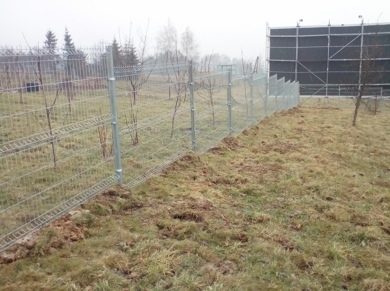 Nowo zainstalowane, ocynkowane, panelowe ogrodzenie z siatki na nierównym terenie w pochmurny dzień, z widocznymi śladami po pracach ziemnych.