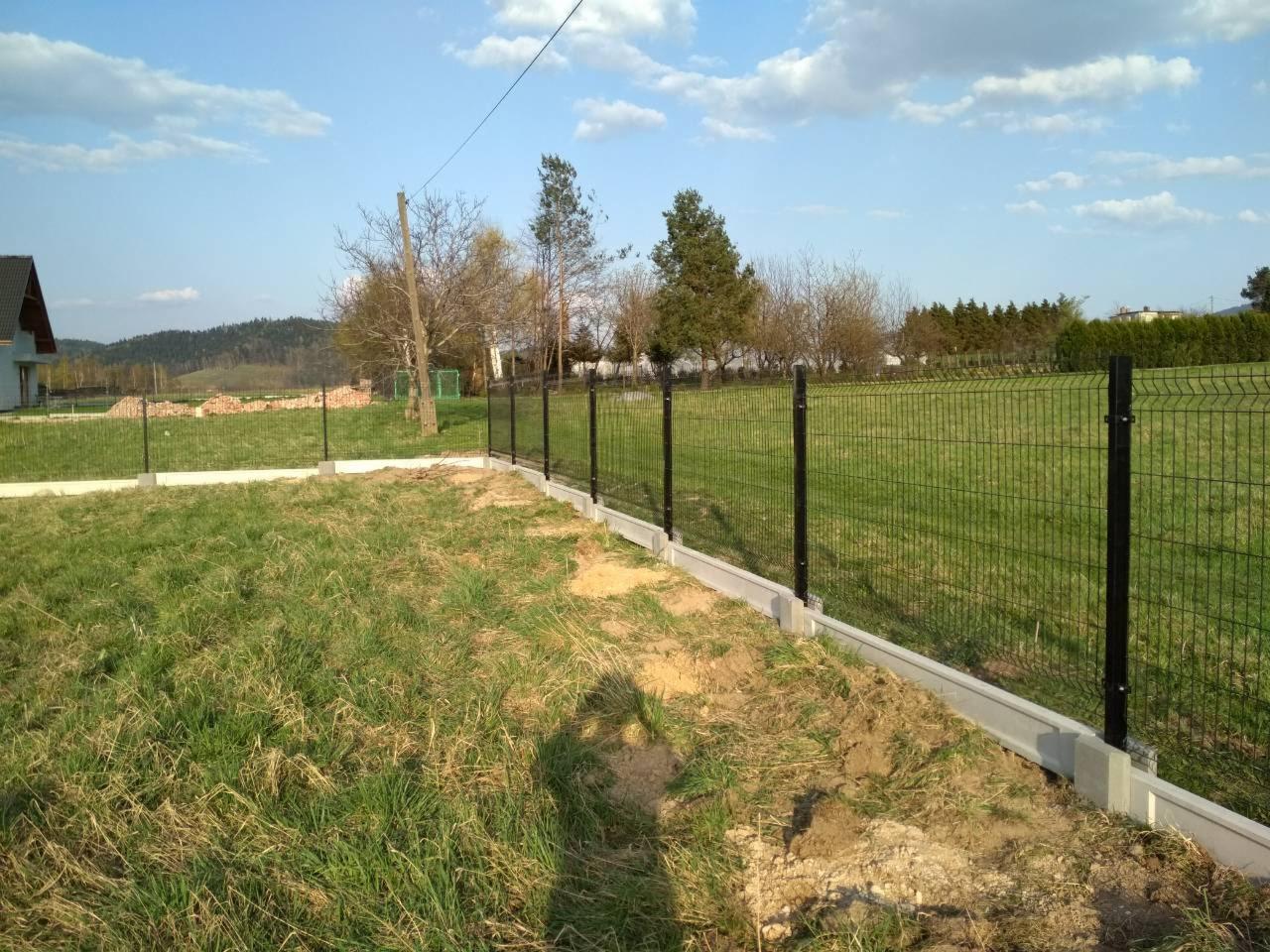 Częściowo ukończony montaż ogrodzenia panelowego z podmurówką betonową na nierównym terenie, widoczne słupki i siatka. Tło: trawa, drzewa i budynek.