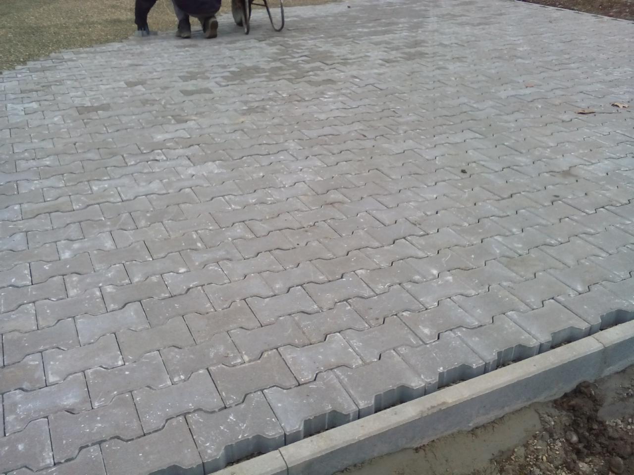 Ułożona szara kostka brukowa na podwórku, widoczne fragmenty nóg robotników i wózek w tle.