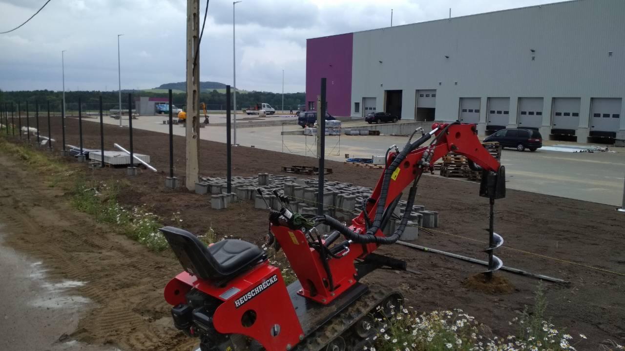 Czerwona wiertnica Heuschrecke przygotowuje teren pod słupki ogrodzeniowe na tle hali magazynowej z rampami załadunkowymi.