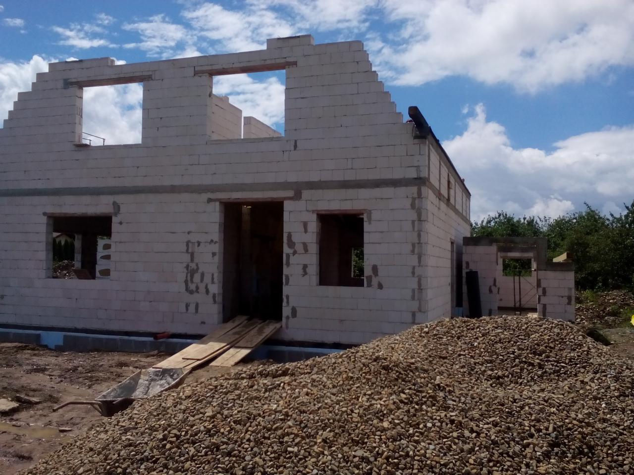 Budowa domu w trakcie wznoszenia murów z białego bloczka, widoczny podjazd z desek i pryzma kamieni na pierwszym planie, błękitne niebo w tle.