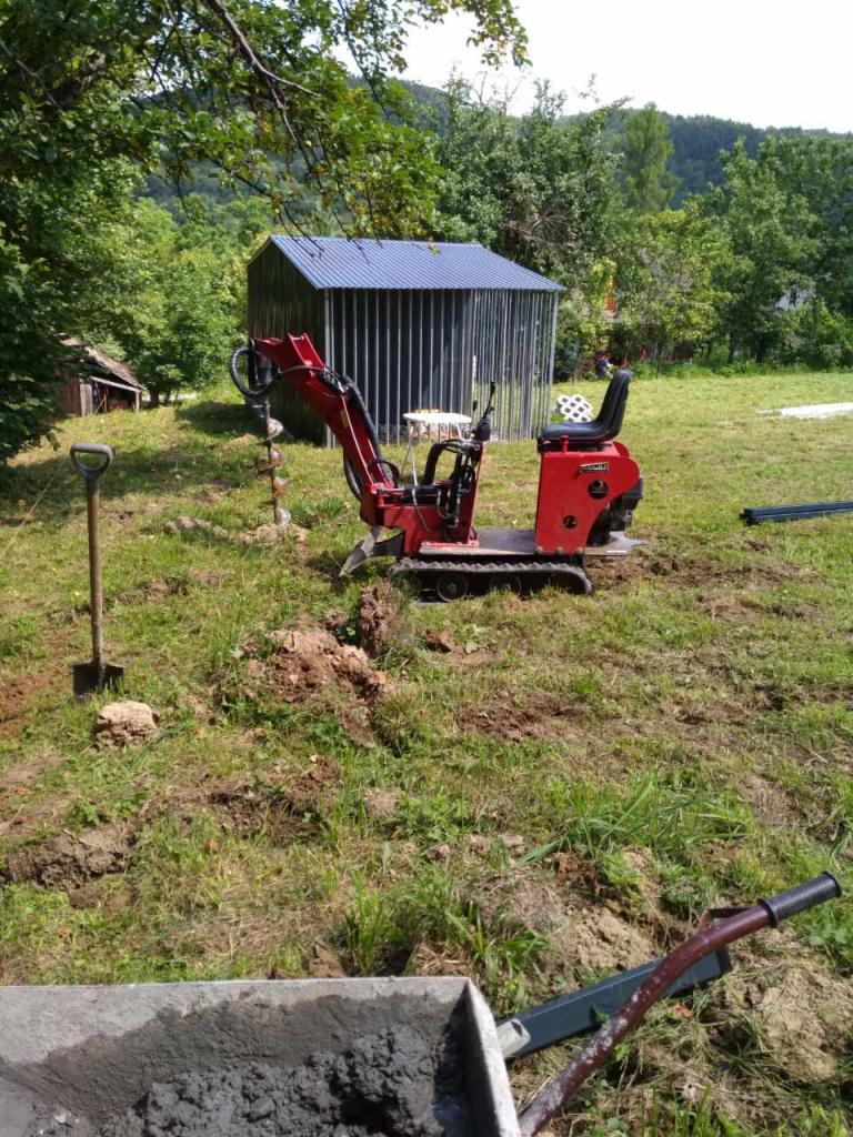 Czerwona mini wiertnica spalinowa wierci otwory pod słupki ogrodzeniowe na trawiastym terenie, widoczna łopata i taczka z betonem, w tle blaszany garaż z niebieskim dachem i zalesione wzgórze.