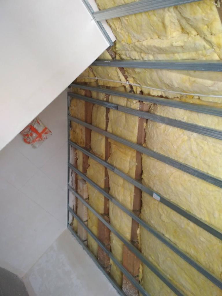 Wnętrze w trakcie ocieplania: żółta wełna mineralna między drewnianymi belkami, stalowa konstrukcja nośna, częściowo zamontowana płyta gipsowo-kartonowa.