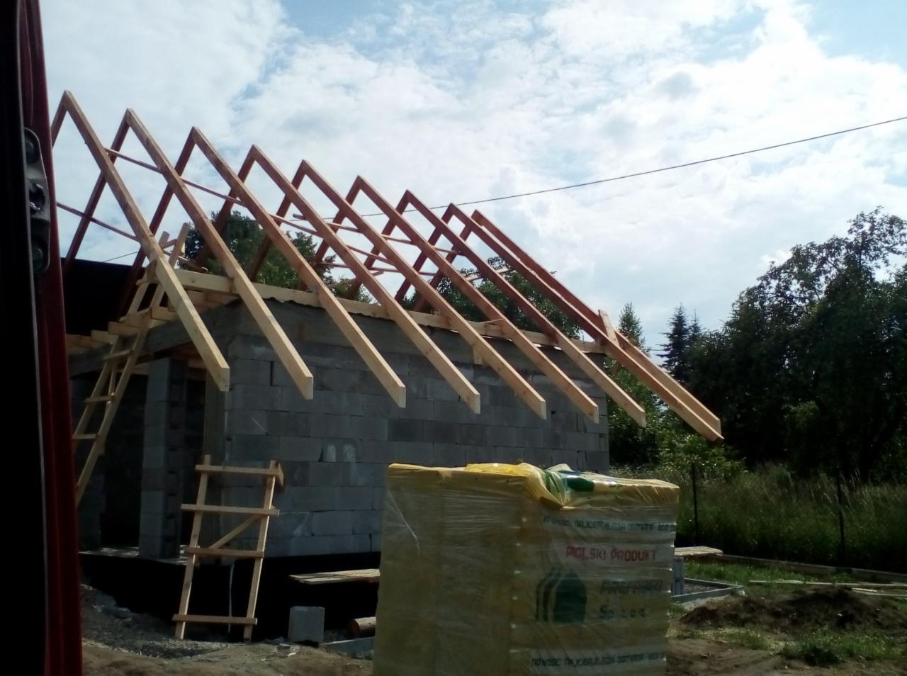 Wznoszenie więźby dachowej na budynku z bloczków betonowych, widoczne drewniane krokwie i dwie drabiny, w tle drzewa i błękitne niebo z chmurami.