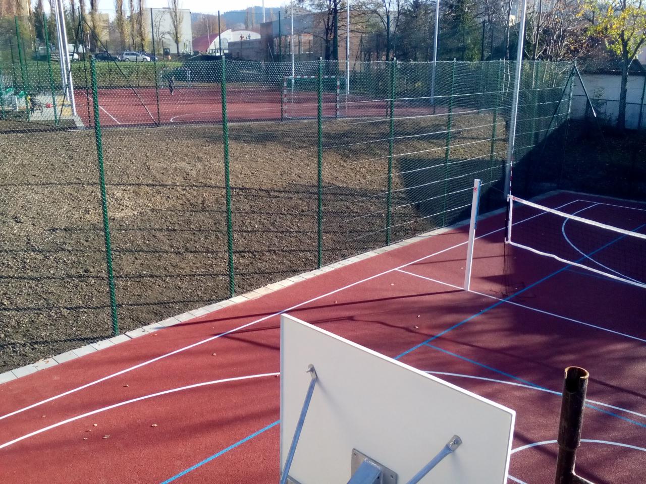 Nowo wybudowane boisko sportowe z czerwoną nawierzchnią, zielonym ogrodzeniem z siatki i elementami wyposażenia do siatkówki i koszykówki, w tle widoczne inne boiska i budynki.