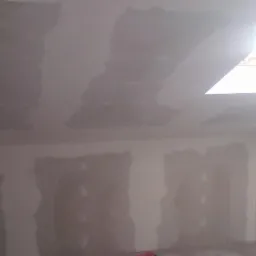 Pokój w trakcie prac przygotowawczych do malowania: ściany i sufit pokryte warstwą szpachli, widoczne nierówności, okno dachowe w suficie wpuszczające jasne światło.