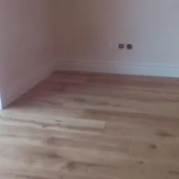 Jasne panele podłogowe ułożone w rogu pomieszczenia, białe listwy przypodłogowe, gładka ściana z widocznymi gniazdkami elektrycznymi.