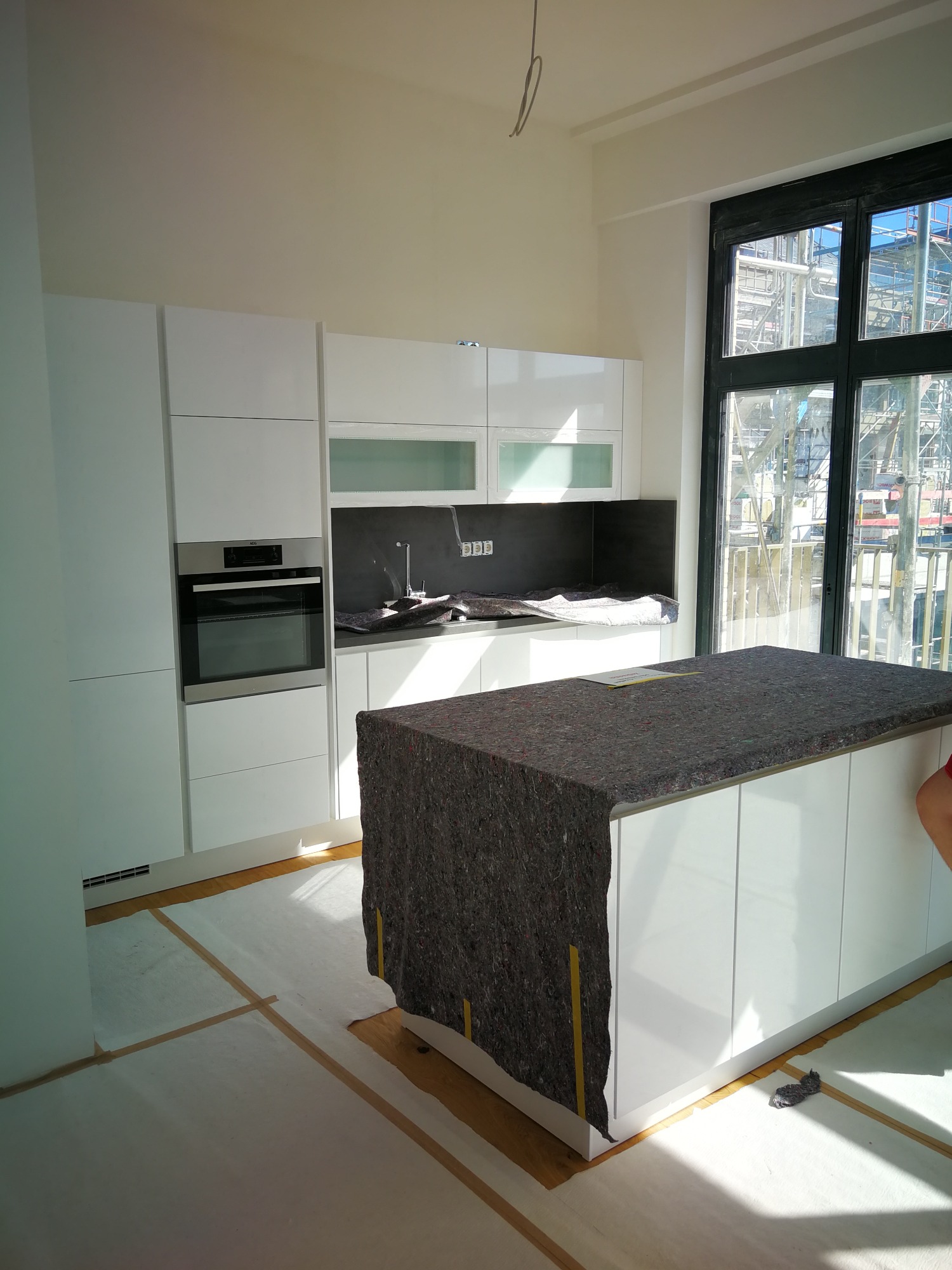 Nowoczesna, biała kuchnia z wyspą, zabezpieczona materiałem ochronnym podczas prac wykończeniowych; widoczny piekarnik marki AEG i okno z widokiem na budowę.