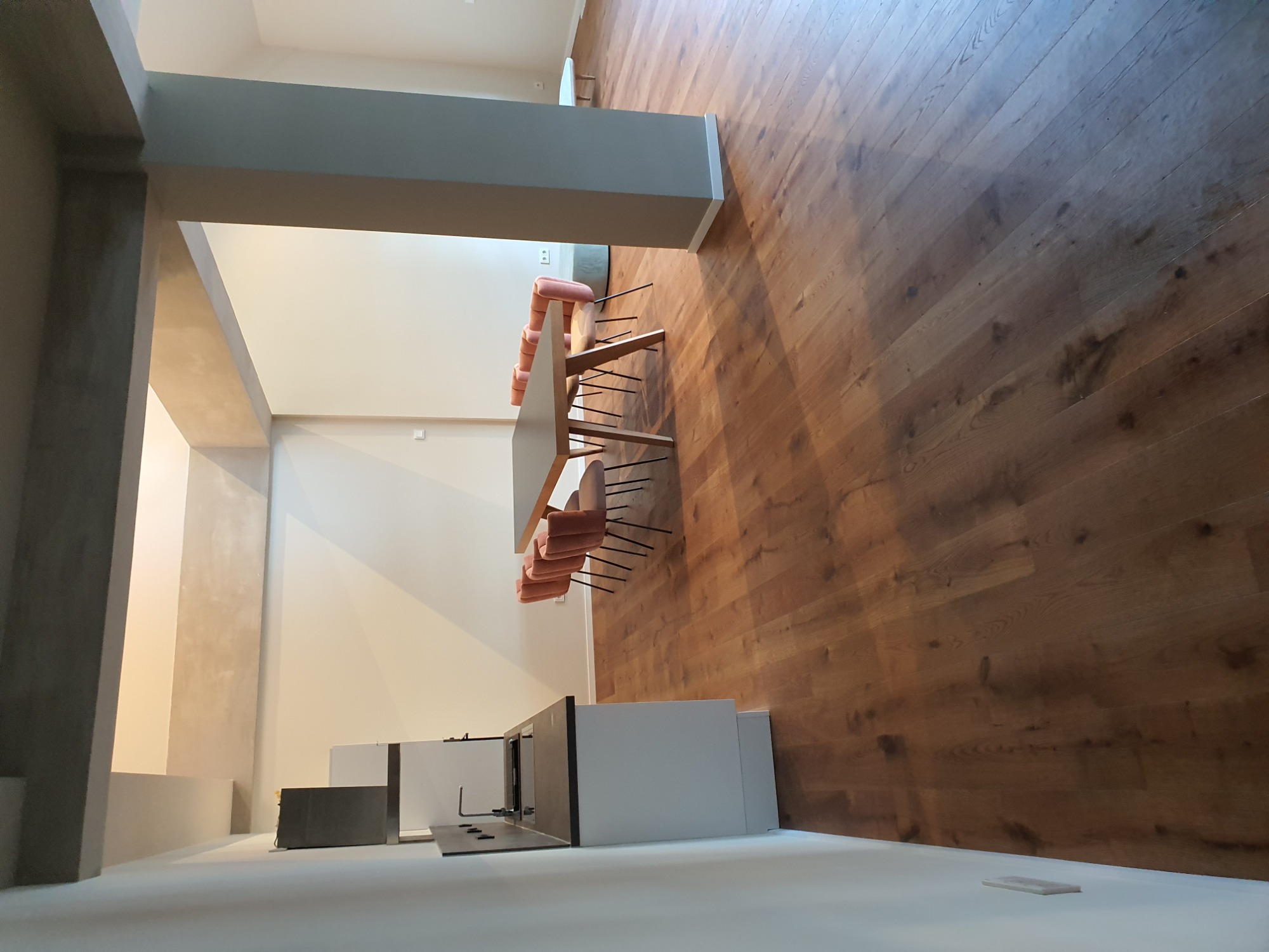 Wysoki kadr na wnętrze w trakcie wykańczania: drewniana podłoga ułożona w jodełkę, surowy betonowy słup, białe ściany i meble kuchenne bez frontów, stół z krzesłami w kolorze pudrowego różu.