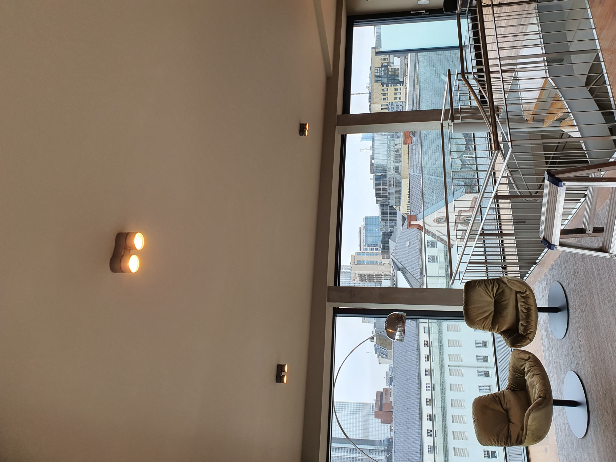 Nowoczesne wnętrze z widokiem na miasto: Dwa krzesła obrotowe, metalowe schody z barierką, oświetlenie ścienne LED i panoramiczne okna.