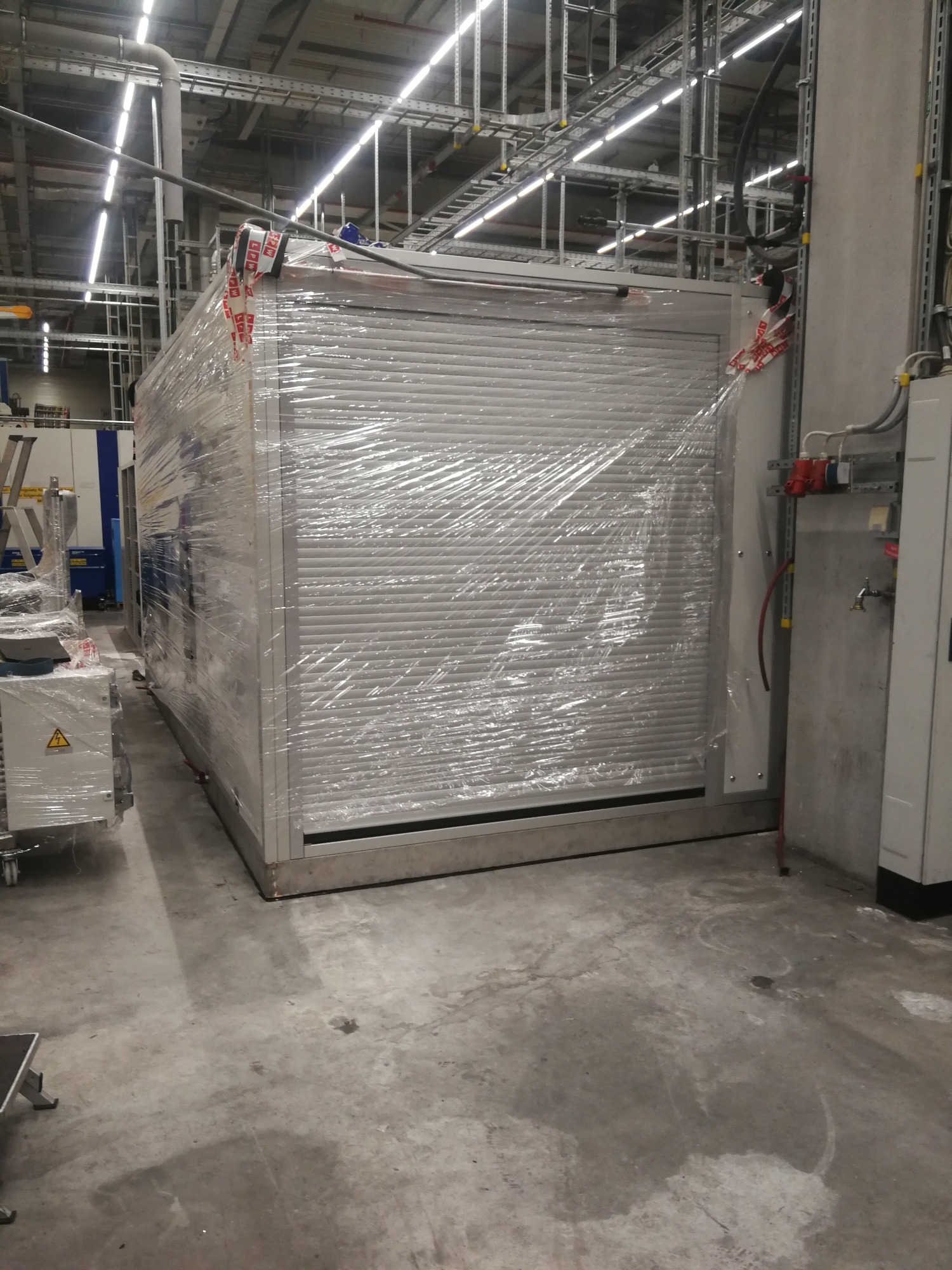 Duża, srebrna szafa z roletą, częściowo owinięta folią ochronną, stoi w hali przemysłowej; widoczne instalacje elektryczne i rury na ścianie w tle.