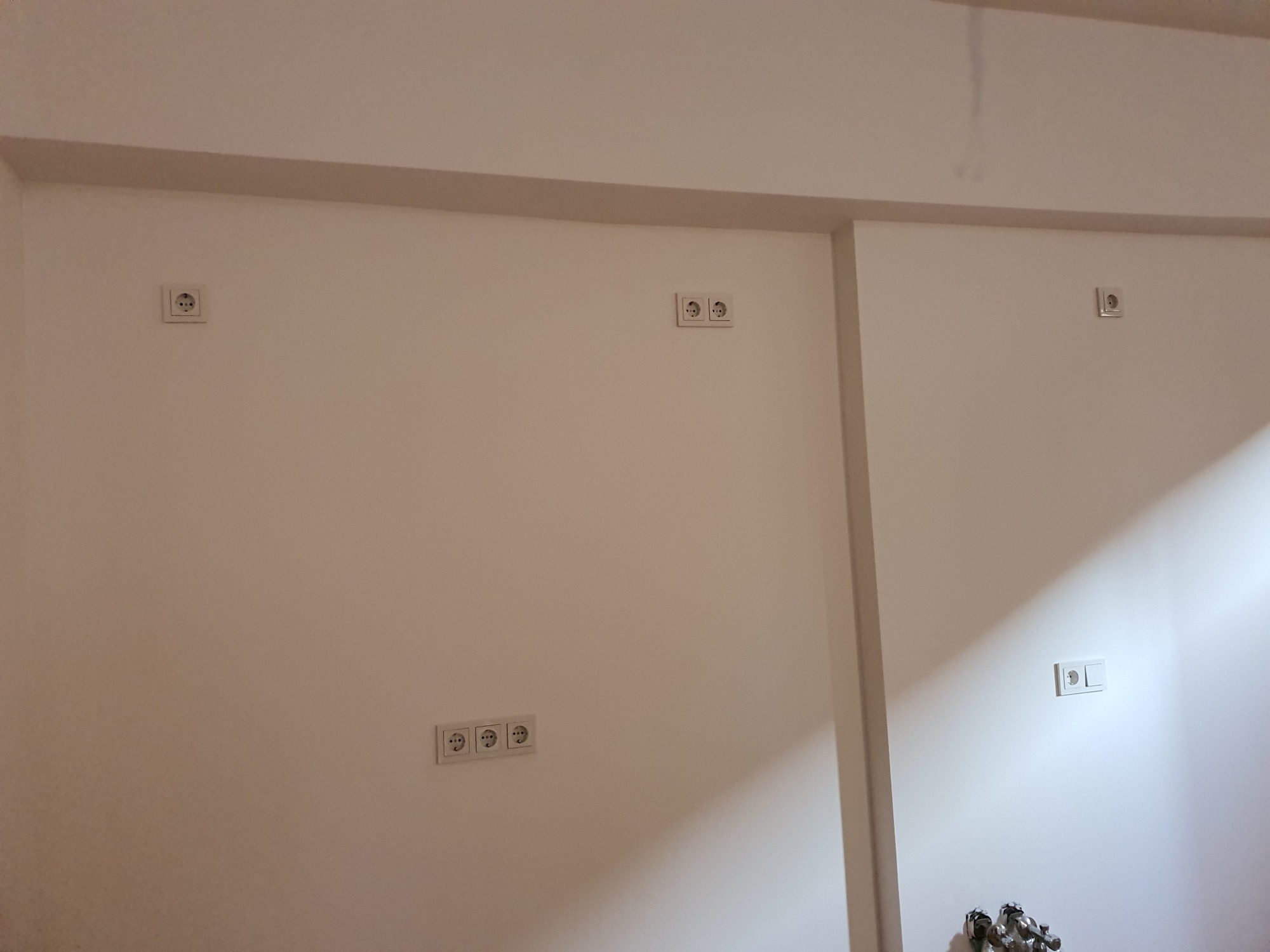 Biała ściana z kilkoma gniazdkami elektrycznymi, niektóre pojedyncze, jedno potrójne, zamontowane na różnych wysokościach.