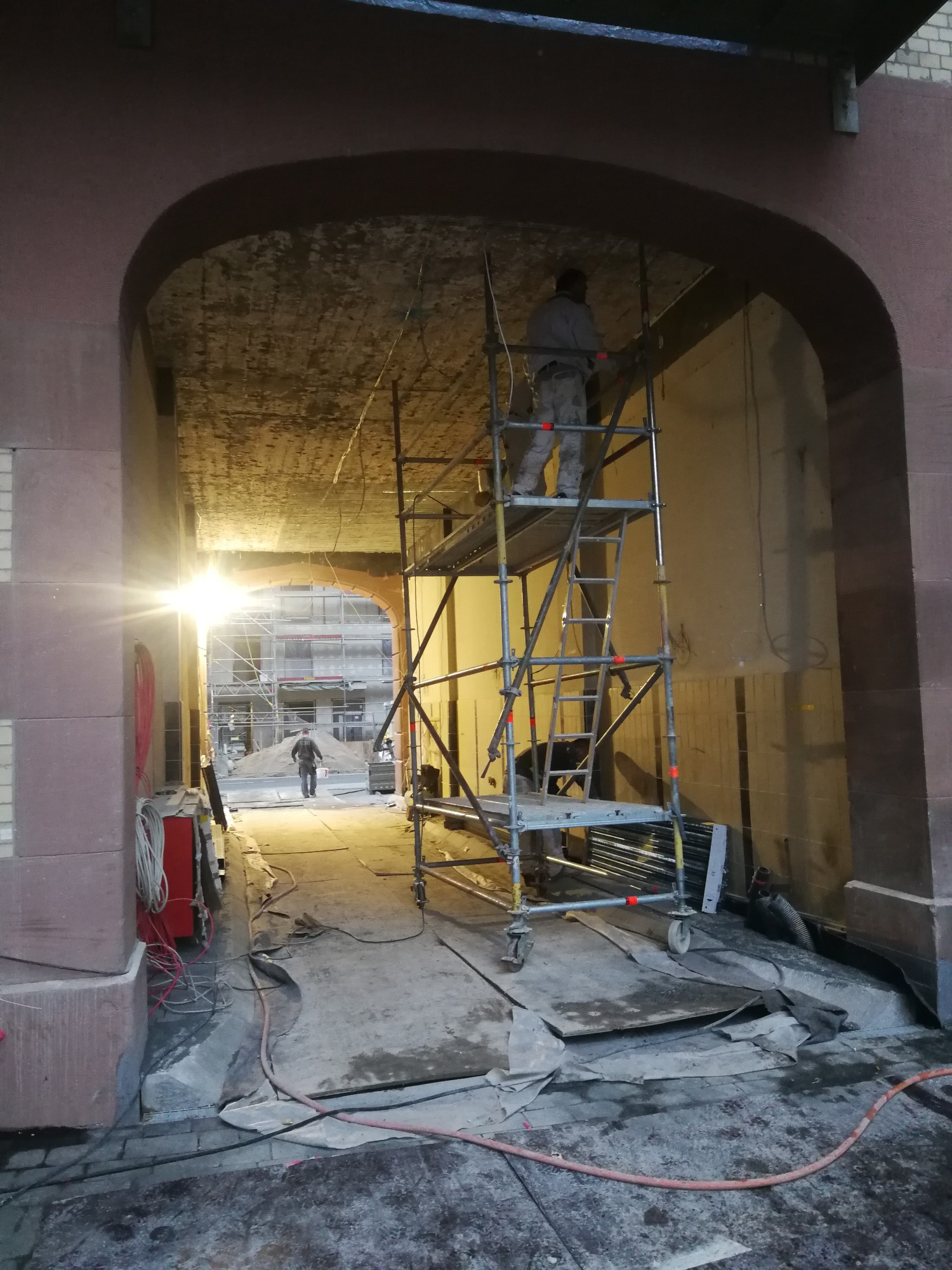 Pracownik na rusztowaniu pod sklepieniem łukowym, instalacja przewodów elektrycznych, widoczne okablowanie i narzędzia w tle.